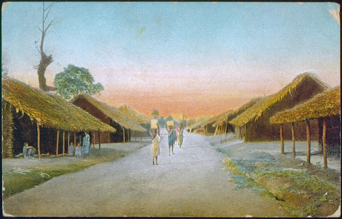 "Händlerdorf in Moschi, Deutsch-Ost-Afrika." ("A traders' village in Moshi, German East Africa."). A road lined with thatched huts. African women are walking on the road and carrying loads on their heads. On the left: an African family (two adults, two children) is standing in front of a hut.; Postcard published by the Leipzig Mission. -- This postcard was sent. -- Cf. original photograph "Strasse in Moschi" ("Road in Moshi"). It can be seen in the Leipzig Mission Archive. -- The Mission station in Moshi was founded in 1896. -- Johannes Schanz (1876-1963) was a missionary of the Leipzig Mission. He served in Moshi and Mamba from 1901 to 1910. Schanz is probably the photographer.